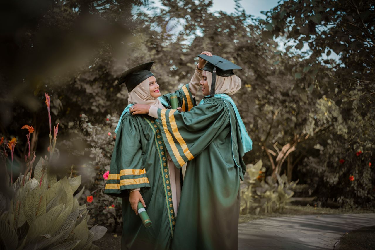 Two women in academic robes celebrate graduation in a lush garden setting.
